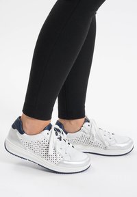 Zapatillas de cuero blancas con detalles en azul marino, diseño perforado, suela plana blanca y cierre de cordones, combinadas con leggings negros.