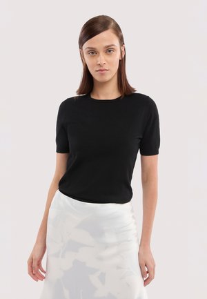 Jeune femme aux cheveux bruns lisses portant un haut noir à manches courtes et une jupe blanche avec un motif abstrait subtil, posant devant un fond blanc.