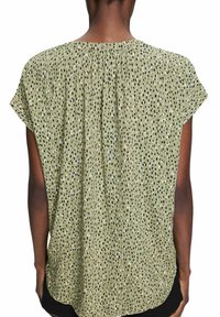 Žena v volné, krátkopsané olivově zelené blůze s černobílým tečkovaným vzorem, viděná zezadu.