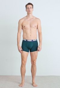 Sötétzöld Puma boxer alsónadrág széles fekete derékkal, amelyen a márkanevet ismétlik. Az anyag simának és rugalmasnak tűnik.