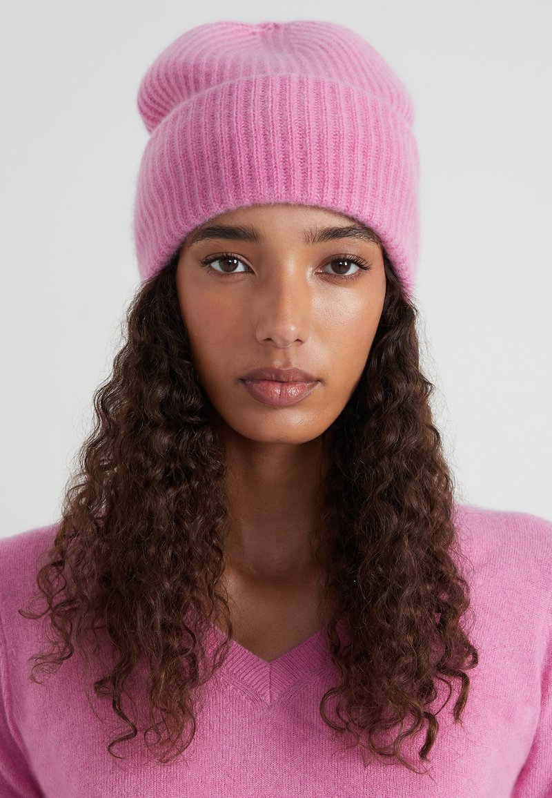 Bonnet tricoté rose avec une texture côtelée. Présente un sommet arrondi et un léger effet dégoulinant. Fabriqué en matériau doux et chaud.