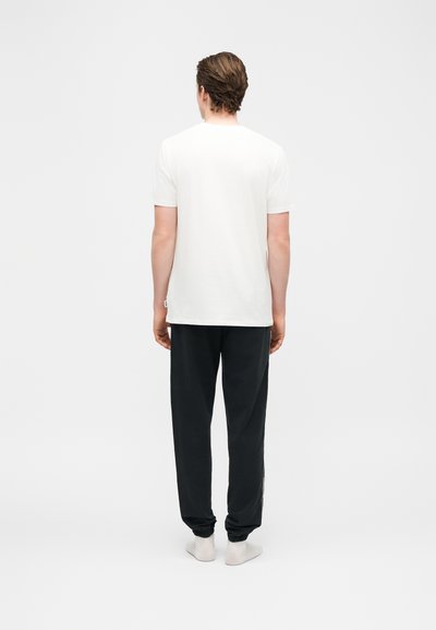 Άνδρας που στέκεται με την πλάτη γυρισμένη, φοράει απλό λευκό κοντομάνικο μπλουζάκι, μαύρο παντελόνι φόρμας και λευκές κάλτσες, μπροστά σε ένα απλό φόντο.