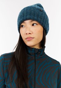 Gorro de punto en azul oscuro con una textura acanalada. Presenta una etiqueta con el logo como acento. Se lleva con una chaqueta de cremallera negra y verde azulado con patrones.