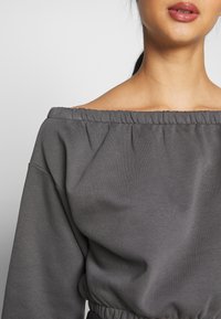 Sudadera gris con los hombros al descubierto, con escote fruncido, mangas largas y cintura ajustada. Tejido suave con una textura sutil.