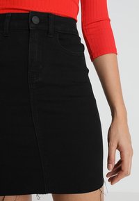 Vestido de mezclilla negro con cintura alta, bolsillos frontales y dobladillo sin terminar. La tela tiene una textura suave con un ligero elastano.