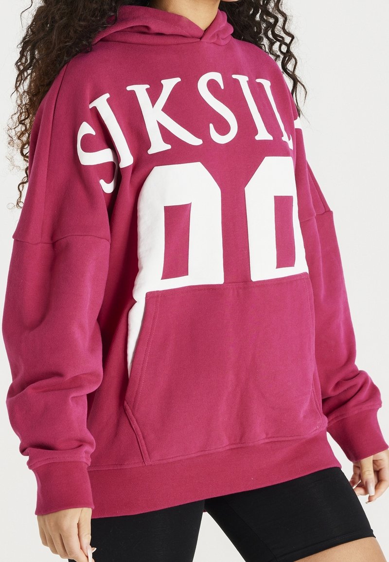 Mujer vestida con un sudadera oversized rosa con el texto blanco "SIKSILK 00" y pantalones cortos negros, de pie frente a un fondo blanco.