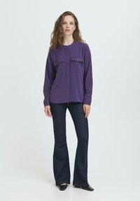 Donna con capelli mossi che indossa una camicia a maniche lunghe viola con bottoni, jeans svasati scuri e scarpe nere a punta, in piedi su uno sfondo bianco.