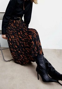 Falda negra con plisados y un patrón abstracto marrón, combinada con botas de tacón alto negras ajustadas. Camisa negra ceñida visible a la altura de la cintura.