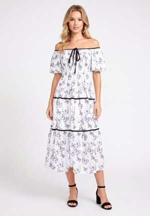 Donna che indossa un vestito bianco scollato con stampa di farfalle nere e tacchi neri con cinturino alla caviglia, in piedi su uno sfondo chiaro e semplice.