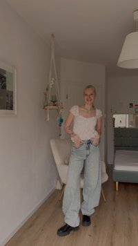 Τοπ από δαντέλα ανοιχτού χρώματος με κοντά μανίκια, ψηλόμεσα ξεθωριασμένα τζιν και μαύρα μποτάκια με πλατφόρμα. Εσωτερικός χώρος σε ουδέτερους τόνους με μια καρέκλα και διακοσμητικά.