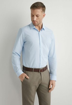 Mężczyzna w jasnoniebieskiej koszuli na guziki i brązowych spodniach z paskiem, stojący przy białej ścianie, patrzący w dół z jedną ręką w kieszeni.