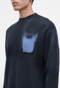 Homme portant un pull côtelé foncé avec une pièce bleu clair présentant un petit logo ovale sur la partie gauche de la poitrine, sur un fond uni.