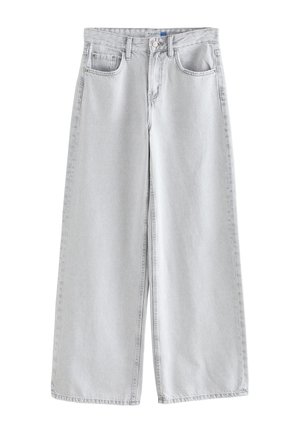 Žena nosi svetlošedé rifle s vysokým pásom, gombíkom a vreckami, skombinované s bielou tielkovou blúzkou a zlatým náramkom.