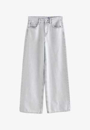 Vrouw draagt lichtgrijze jeans met hoge taille, knoop en zakken, gecombineerd met een witte mouwloze blouse en een gouden armband.