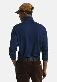 Marineblauwe trui met een hoge kraag en geribbelde manchetten, gecombineerd met bruine corduroy broek en een lichtbruine geruite pet met een donkere rand.