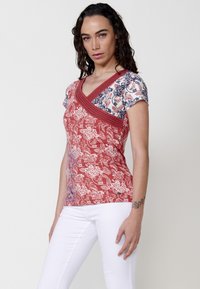 Kurzes rotes Oberteil mit floralem Muster, einem Kreuzkragen und kontrastierenden Schultern in blau-weißem Muster. Weicher Stoff, figurbetontes Design.
