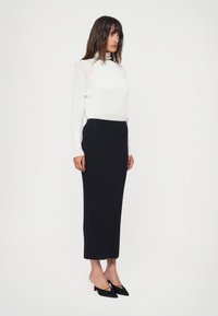Un top alb cu mansete lungi, texturat cu nervuri, asortat cu o fustă midi neagră, texturată cu nervuri. Modelul poartă pantofi cu toc ascuțit negri. Fundal simplu.