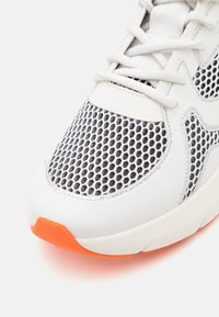 Gros plan d'une basket blanche avec tige en mesh noir, lacets blancs et bout orange sur une semelle blanche.
