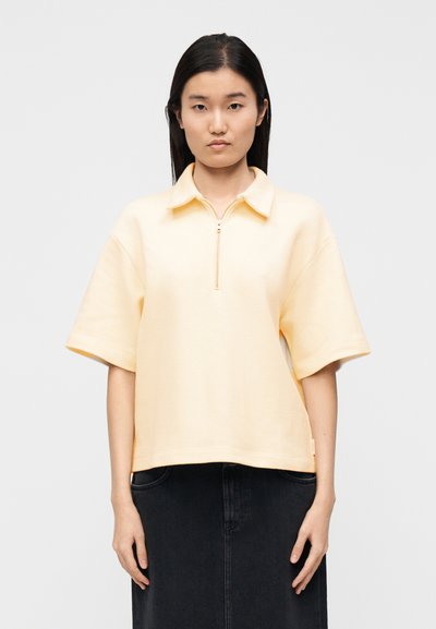 Pull-over jaune à manches courtes avec un col zippé, en tissu texturé. Il présente un col et une coupe décontractée, associé à une jupe en denim foncé.