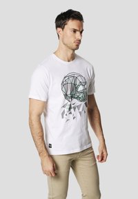 T-shirt en coton blanc avec un motif géométrique de crâne en vert et noir, orné de motifs triangulaires. Manches courtes unies, col rond.