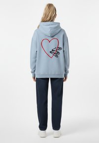 Felpa blu cielo con un cuore rosso e spille di sicurezza nere ricamate sul retro, abbinata a pantaloni scuri a fantasia e scarpe bianche.