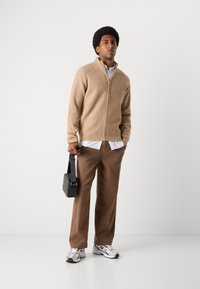 Beige zip-up genser med ribbestruktur, hvitstripet skjorte under, brune bukser, svart crossbody veske og grå joggesko.