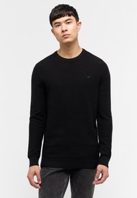 Schwarzer Strickpullover mit Rundhalsausschnitt, langen Ärmeln und gerippten Säumen. Besitzt ein kleines Logo auf der Brust. Wird mit dunklen Jeans getragen.