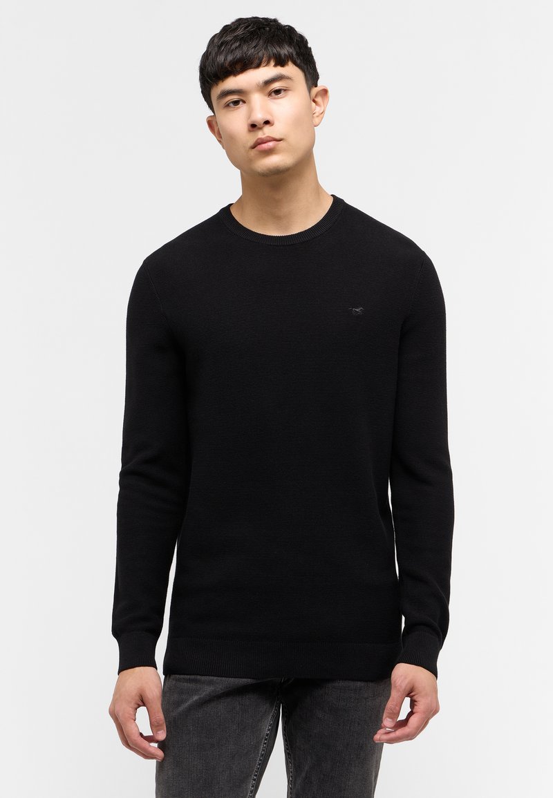 Schwarzer Strickpullover mit Rundhalsausschnitt, langen Ärmeln und gerippten Säumen. Besitzt ein kleines Logo auf der Brust. Wird mit dunklen Jeans getragen.