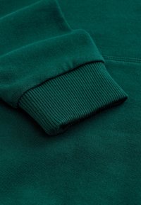 Tissu de sweat-shirt vert foncé avec des détails de poignet côtelés. Texture douce avec une finition lisse. Design simple et épuré sans motifs visibles.