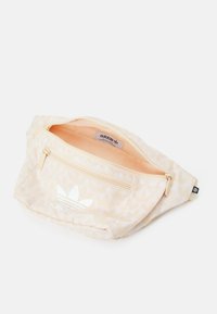 Ljusbeige Adidas midjeväska med vitt trefoil-logotyp, framficka med dragkedja och delvis öppen huvudfack på vit bakgrund.