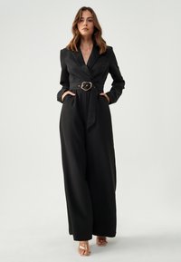 Zwarte jumpsuit met een getailleerde taille, lange mouwen en een klassieke kraag. Voorzien van een gouden riem, twee zijzakken en een wijde broekspijp.