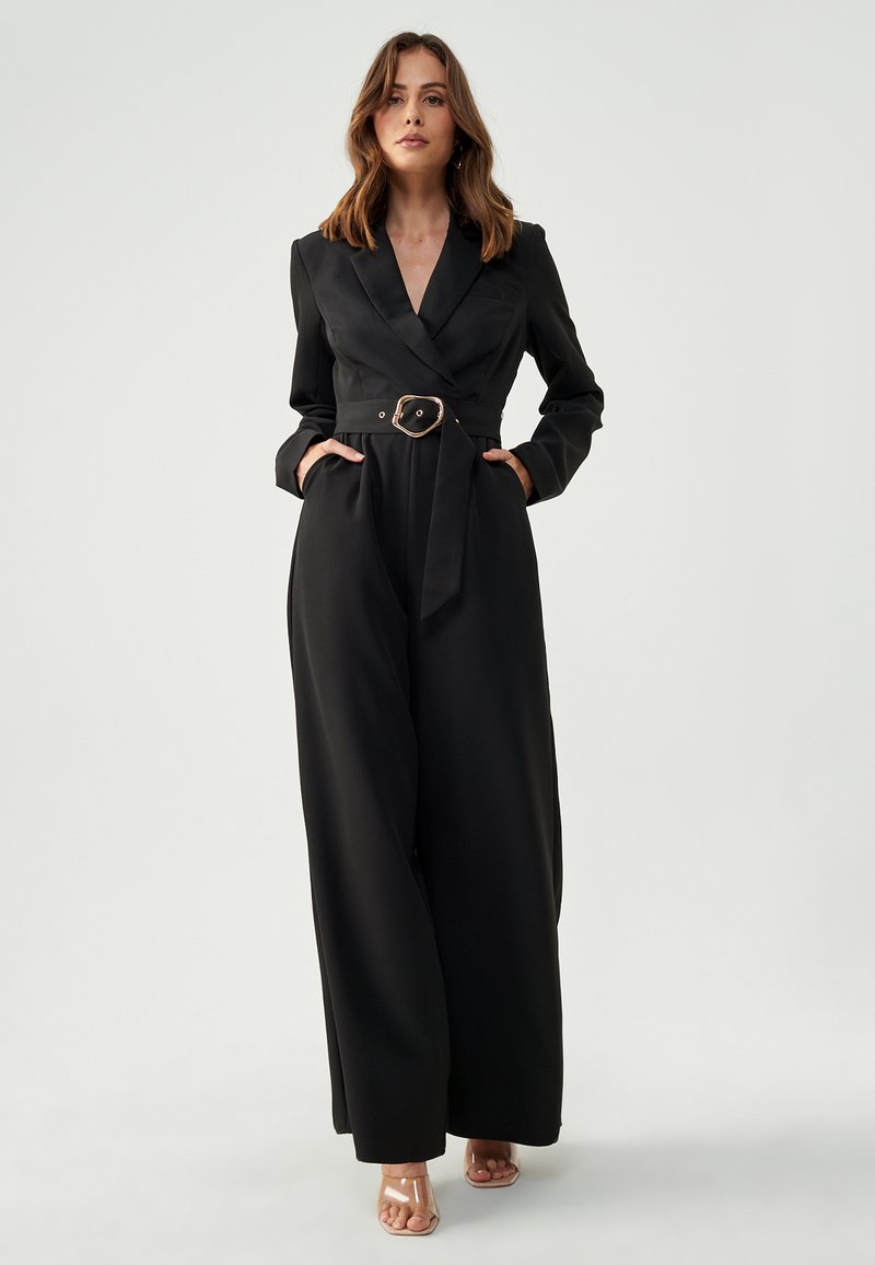 Zwarte jumpsuit met een getailleerde taille, lange mouwen en een klassieke kraag. Voorzien van een gouden riem, twee zijzakken en een wijde broekspijp.