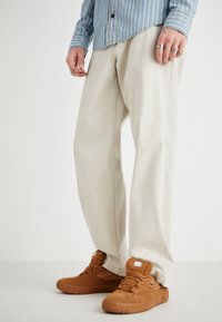 Pantaloni beige a gamba larga in cotone, abbinati a sneakers in camoscio marrone con logo bianco. La camicia è blu con righe bianche.