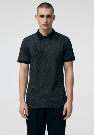 Młody mężczyzna ubrany w czarną, krótkorękawową koszulkę polo z wzorem i kołnierzykiem, stojący na tle jasnego, jednolitego tła.