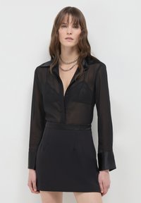 Frau trägt eine transparente schwarze Bluse mit einem Spitzen-BH darunter, kombiniert mit einem schwarzen Rock und geschichteten silbernen Kettenhalsketten, stehend vor einem weißen Hintergrund.