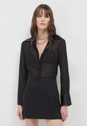 Frau trägt eine transparente schwarze Bluse mit einem Spitzen-BH darunter, kombiniert mit einem schwarzen Rock und geschichteten silbernen Kettenhalsketten, stehend vor einem weißen Hintergrund.