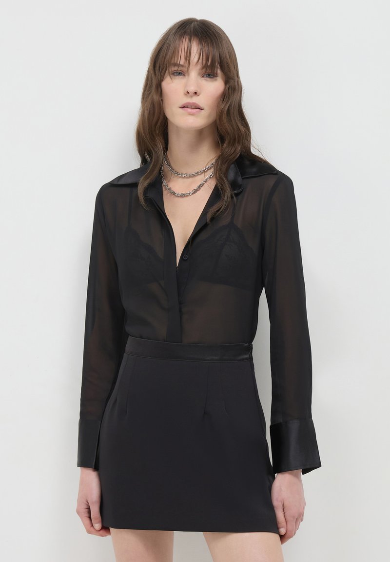 Frau trägt eine transparente schwarze Bluse mit einem Spitzen-BH darunter, kombiniert mit einem schwarzen Rock und geschichteten silbernen Kettenhalsketten, stehend vor einem weißen Hintergrund.