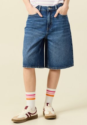 Persoon draagt knielange blauwe spijkershorts, witte sokken met roze en oranje strepen, en beige sneakers met bordeauxrode details.