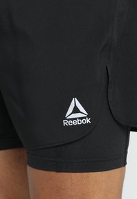 Černé atletické šortky vyrobené z lehkého, pružného materiálu. Vybaveny bílým logem Reebok. Obsahují boční rozparky pro větší pohyblivost a pohodlí.