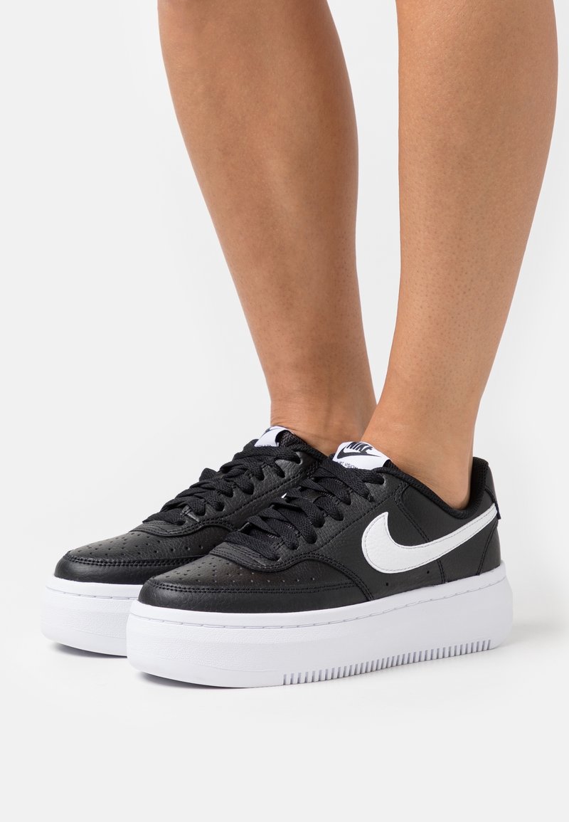 Baskets noires Nike avec logo swoosh blanc et semelles blanches épaisses, portées pieds nus sur un fond clair uni.