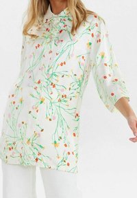 Femme portant une blouse blanche à manches longues avec un motif floral vert, orange, jaune et rose, associée à un pantalon blanc sur fond uni.