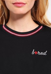 Inquadratura ravvicinata di una donna che indossa una maglietta nera con un colletto a righe rosse e bianche e la parola "bored" ricamata con una forma di cuore rossa.