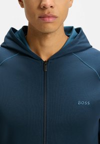 Felpa con zip blu navy con finitura testurizzata, cappuccio interno a contrasto e logo "BOSS" sul petto sinistro. Presenta un design aderente e chiusura con zip.