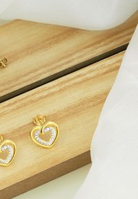 Orecchini a forma di cuore in oro con intarsio di cristallo, poggiati su una superficie di legno accanto a un tessuto bianco. Il design presenta un accento a forma di anello.