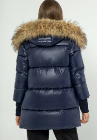 Marineblauwe gewatteerde jas met een nepbontcapuchon. Heeft een glanzende textuur en een horizontaal geborduurd patroon. De lengte reikt tot de dijen en heeft driekwartmouwen.