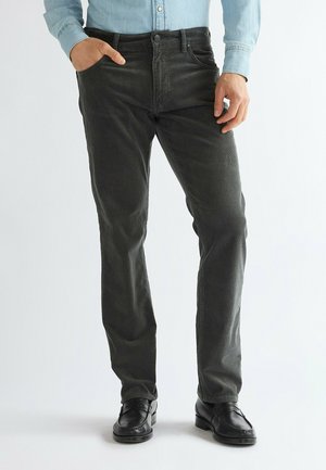 Uomo che indossa pantaloni di velluto grigio scuro, camicia azzurra con bottoni e scarpe eleganti nere con le mani nelle tasche, in piedi su sfondo bianco.