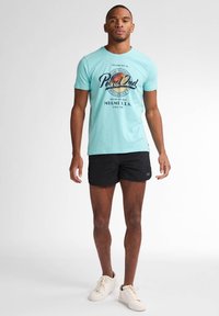 Tricou turcoaz cu maneci scurte, cu imprimeu grafic circular, asortat cu pantaloni scurți negri. Purtați cu adidași albi, stând pe un fundal simplu.