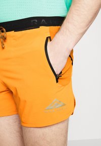 Gros plan d'une personne portant un short Nike Trail orange avec une main dans la poche latérale zippée et une chemise verte rentrée.