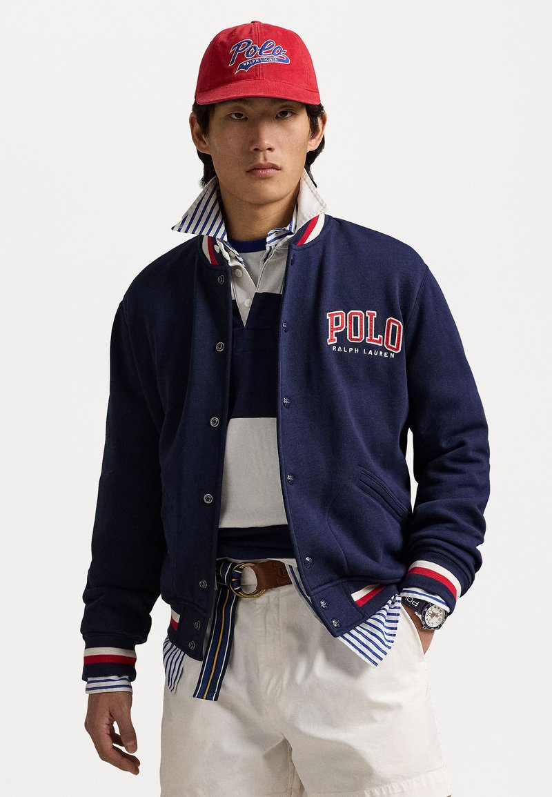 Casaco varsity azul-marinho com detalhes em vermelho e branco, apresentando o logótipo "Polo RALPH LAUREN", gola listrada, combinado com uma polo cinza e calções bege.