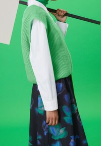 Grön stickad väst över en vit skjorta med knappar, kombinerat med en mörkblommig kjol. Bakgrunden är en enfärgad grön.